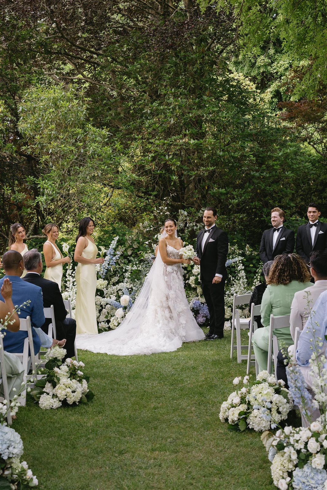 A couple smiling at their garden ceremony with their bridal party, the aisle lined with white and blue hydrangeas