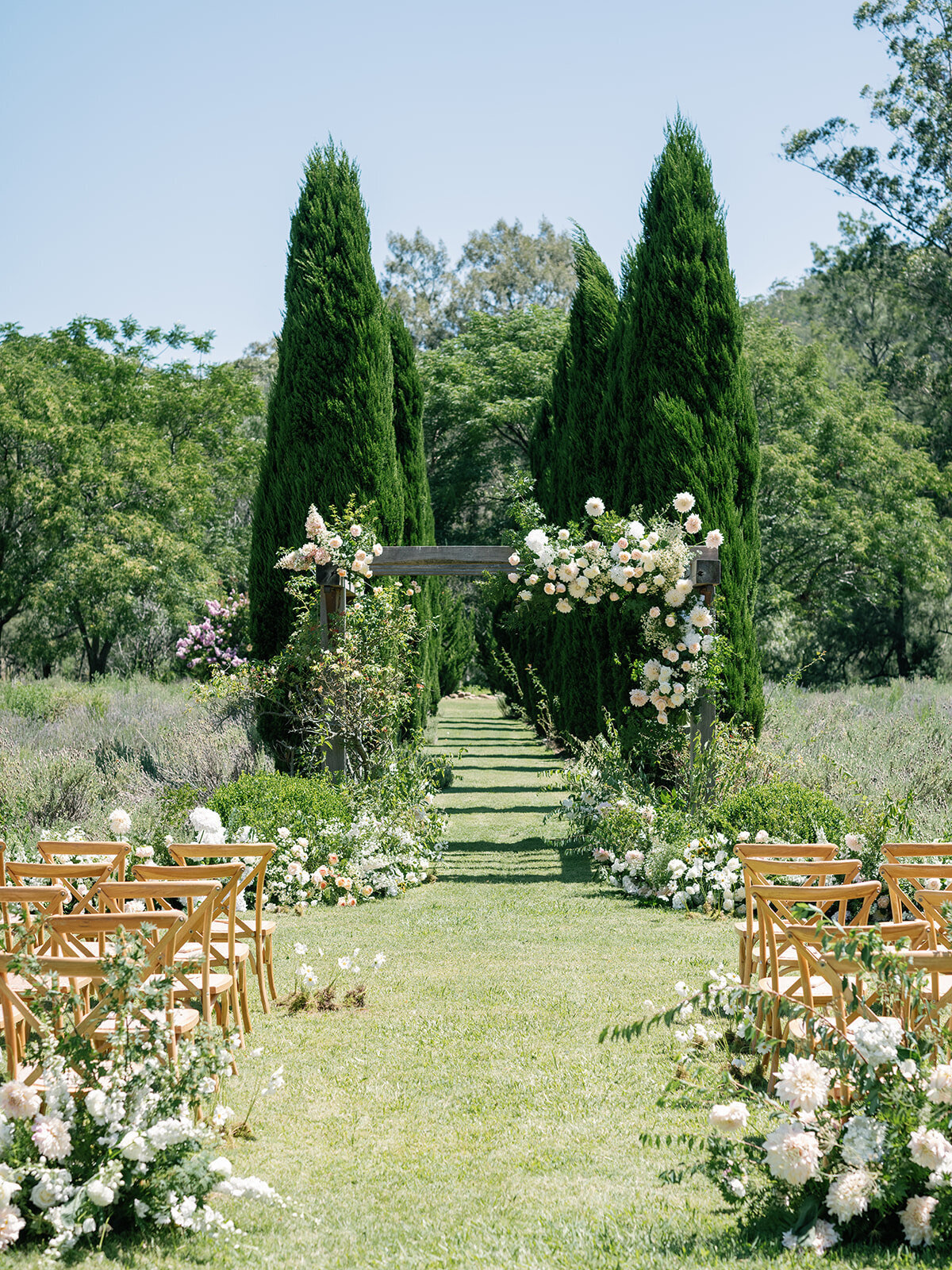 A garden ceremony aisle lined with white flowers between tall cypress trees