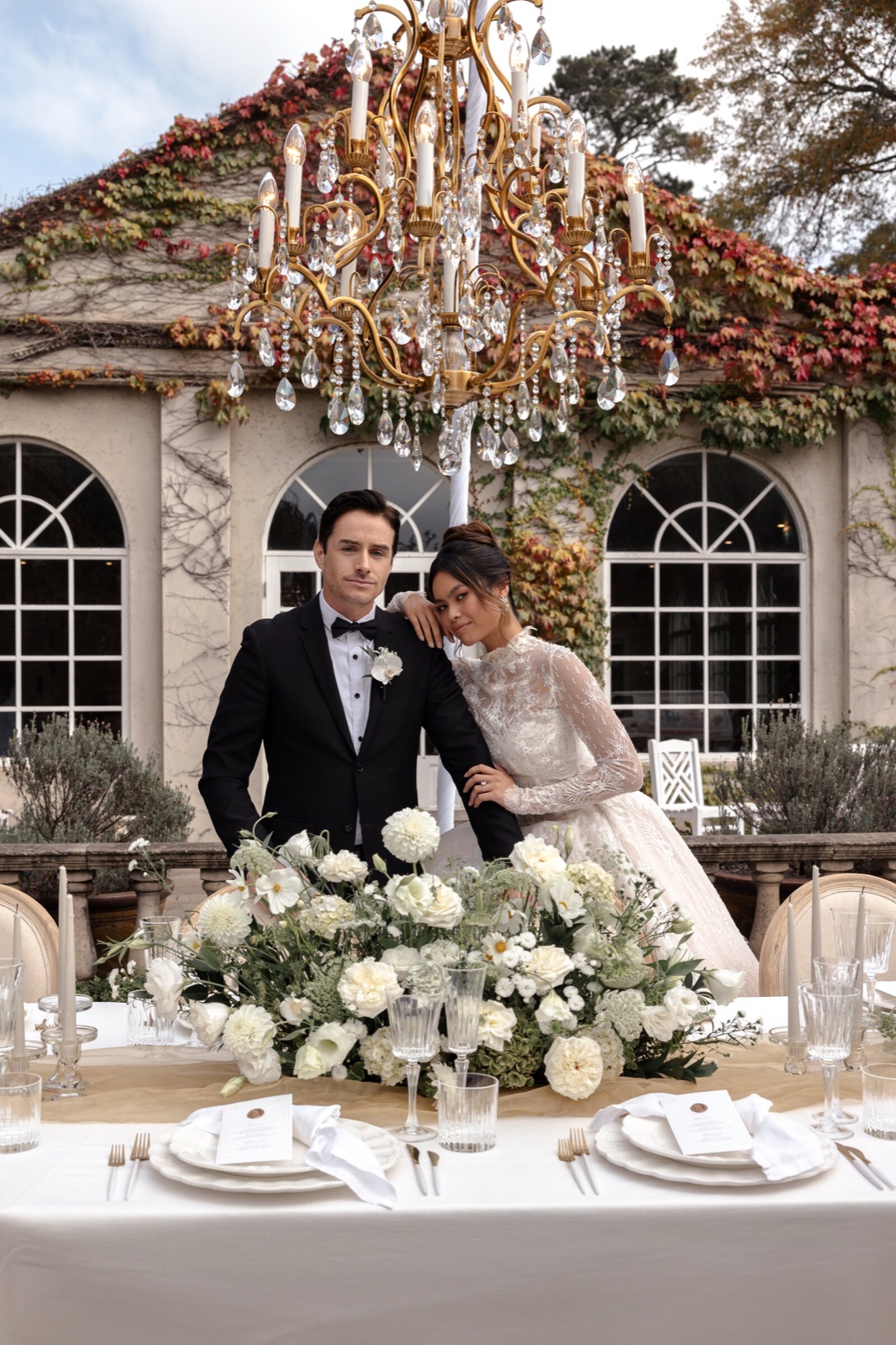 A couple behind their reception table under a gold chandelier outside an arched orangery