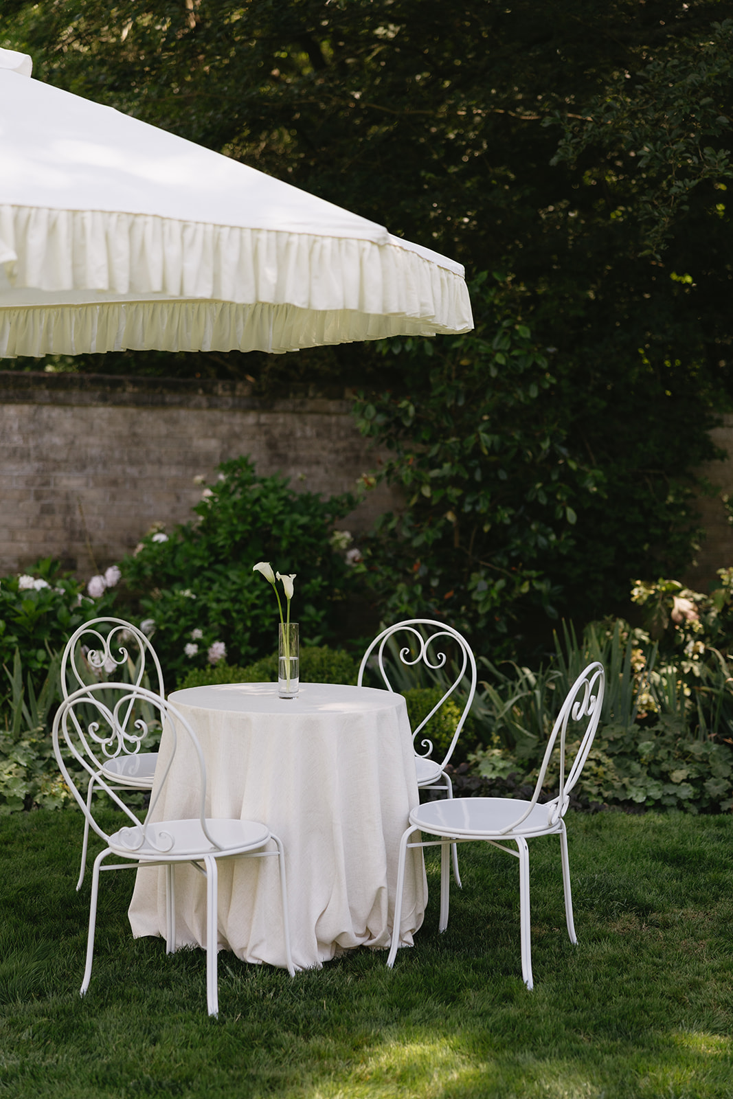 A linen-draped round table with a single calla lily and white heart-back chairs under a ruffled umbrella in a walled garden