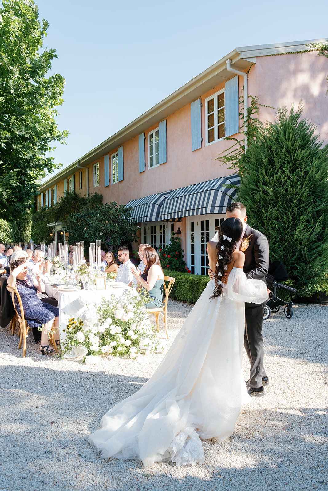 A couple kissing beside a long dinner table of guests outside a pink house