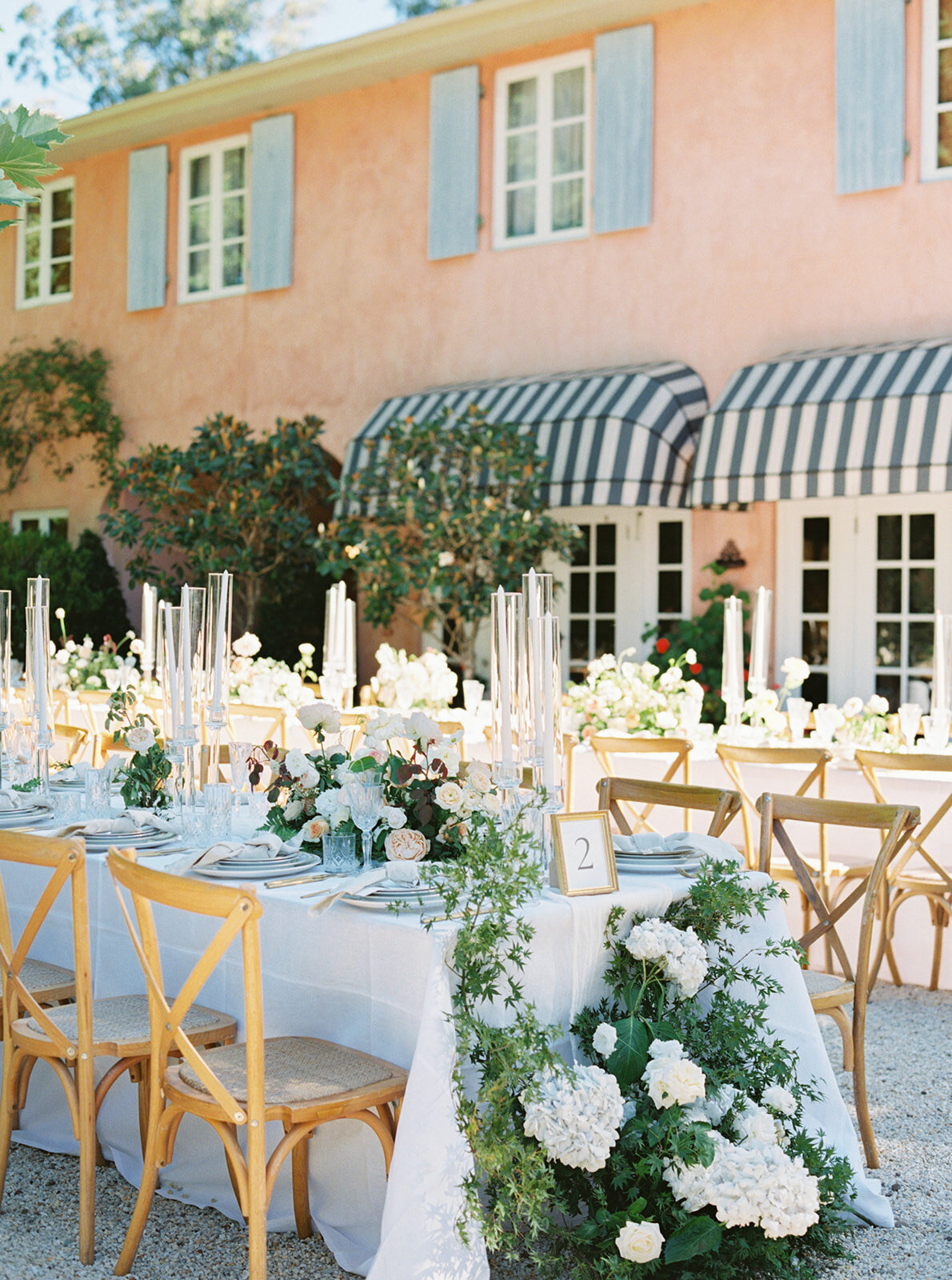 A long outdoor table set with candles and white flowers in front of a pink Italian-style house