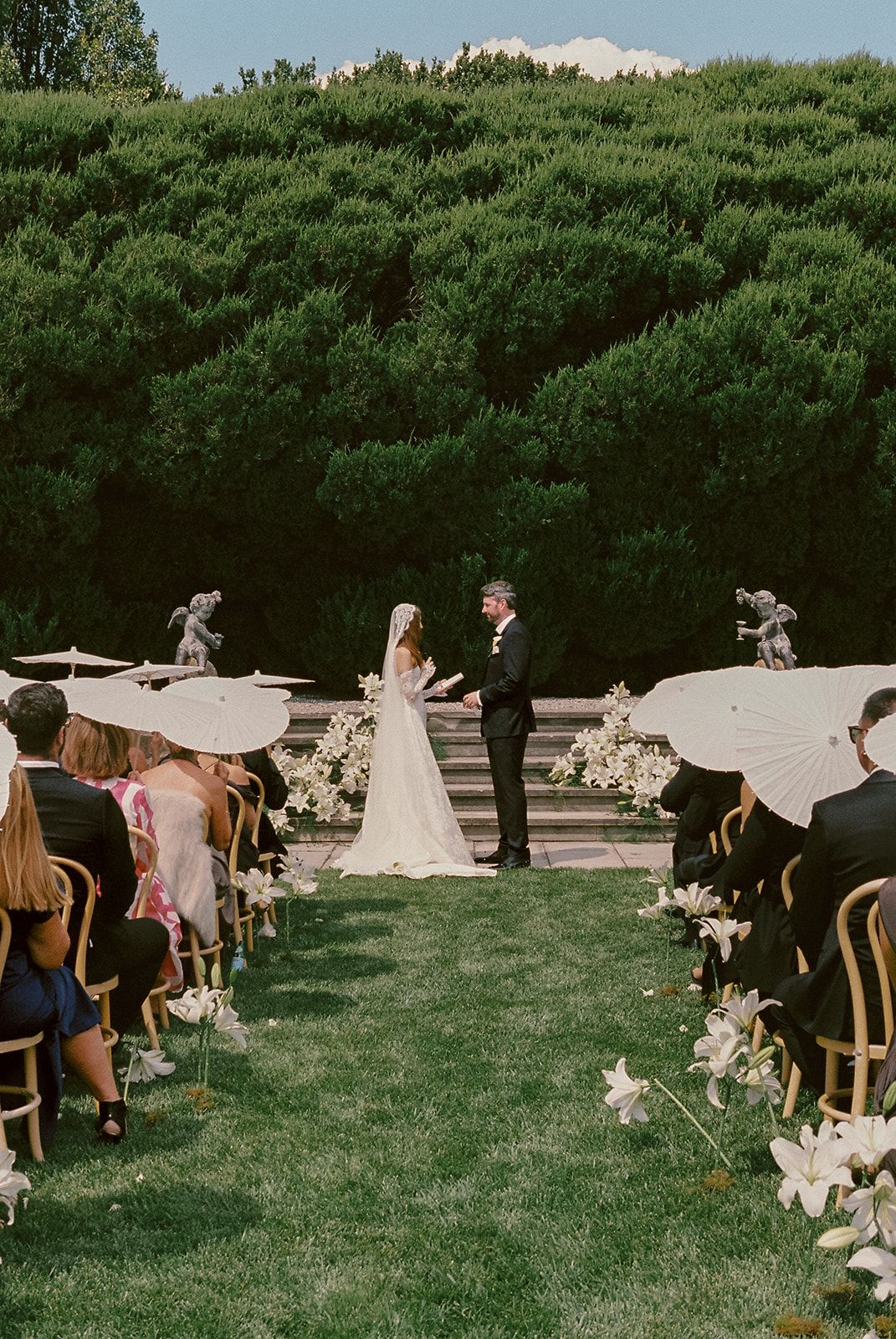 A couple exchanging vows on lily-lined stone steps before a tall hedge, guests holding white parasols