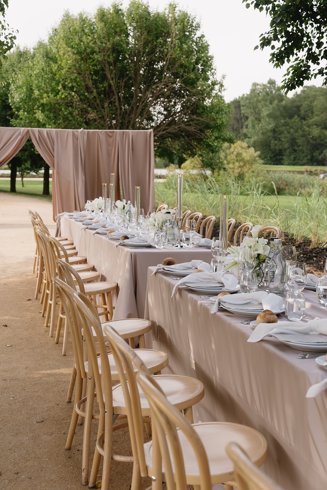 Long tables in blush linen with white calla lilies and tall candles, a draped fabric backdrop behind them in a garden